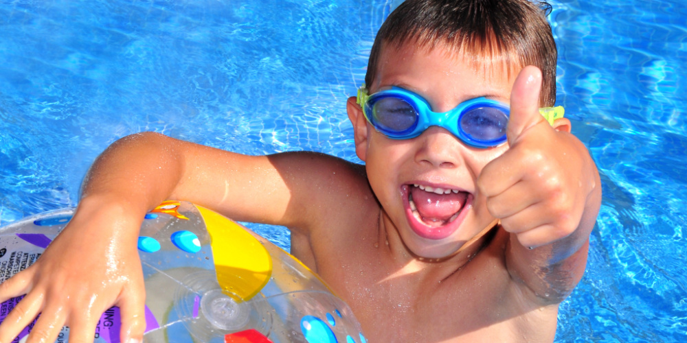 Ein lächelnder Junge mit blauer Schwimmbrille hält einen Wasserball im Schwimmbecken und zeigt den Daumen nach oben.